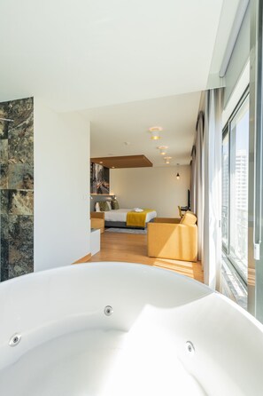 Habitación de hotel moderna con una bañera grande, una cama con ropa de cama amarilla y vistas a la ciudad a través de amplios ventanales.