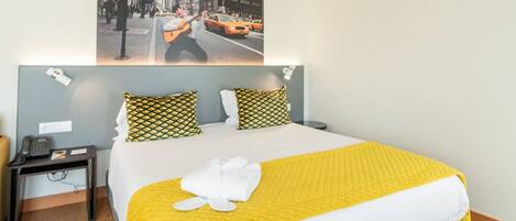 A modern hotel room with a large bed, a headboard with a cityscape photo, a bedside table with a lamp, and a small desk with a phone.