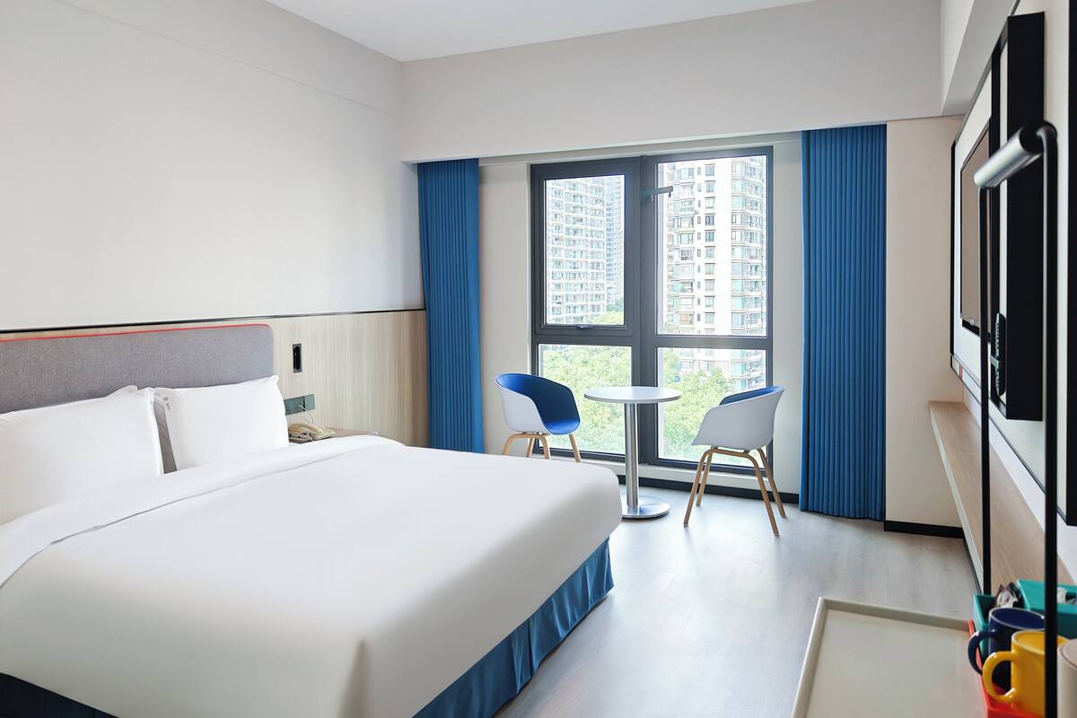 A hotel room with a large bed, two chairs, a small table, a TV, and a window with a city view.