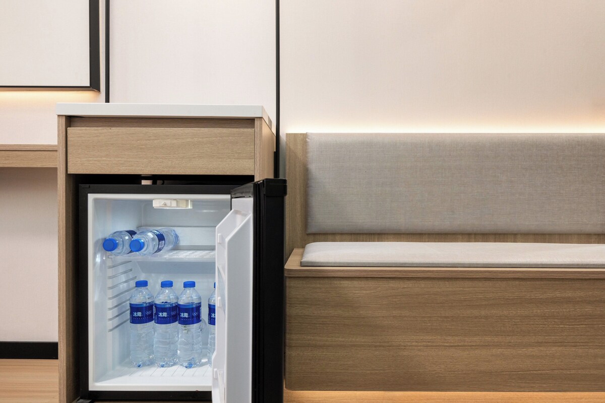 A compact refrigerator with water bottles inside, positioned next to a wooden bedside table with a light-colored cushion.