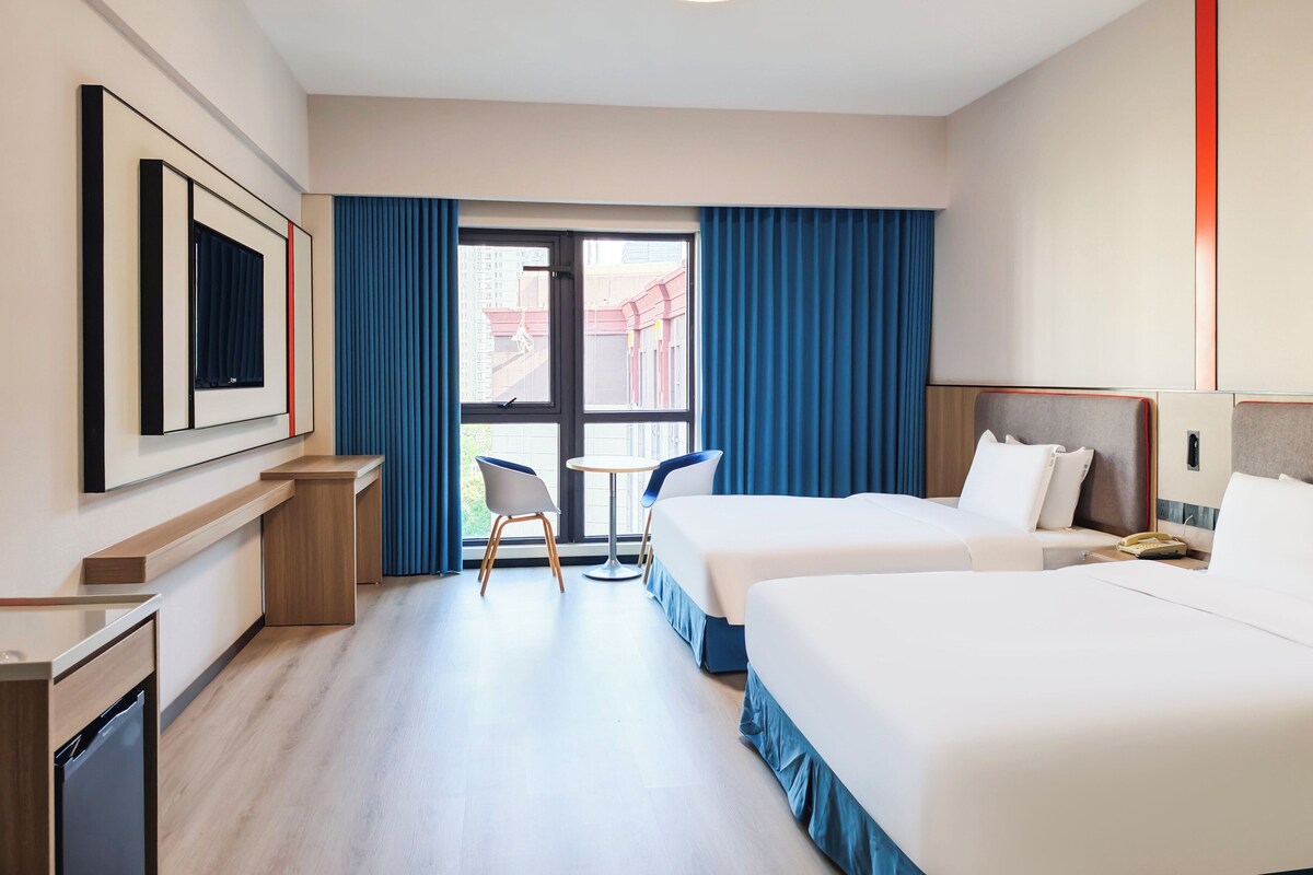 A hotel room with two beds, a desk, a chair, a TV, and a window with curtains.