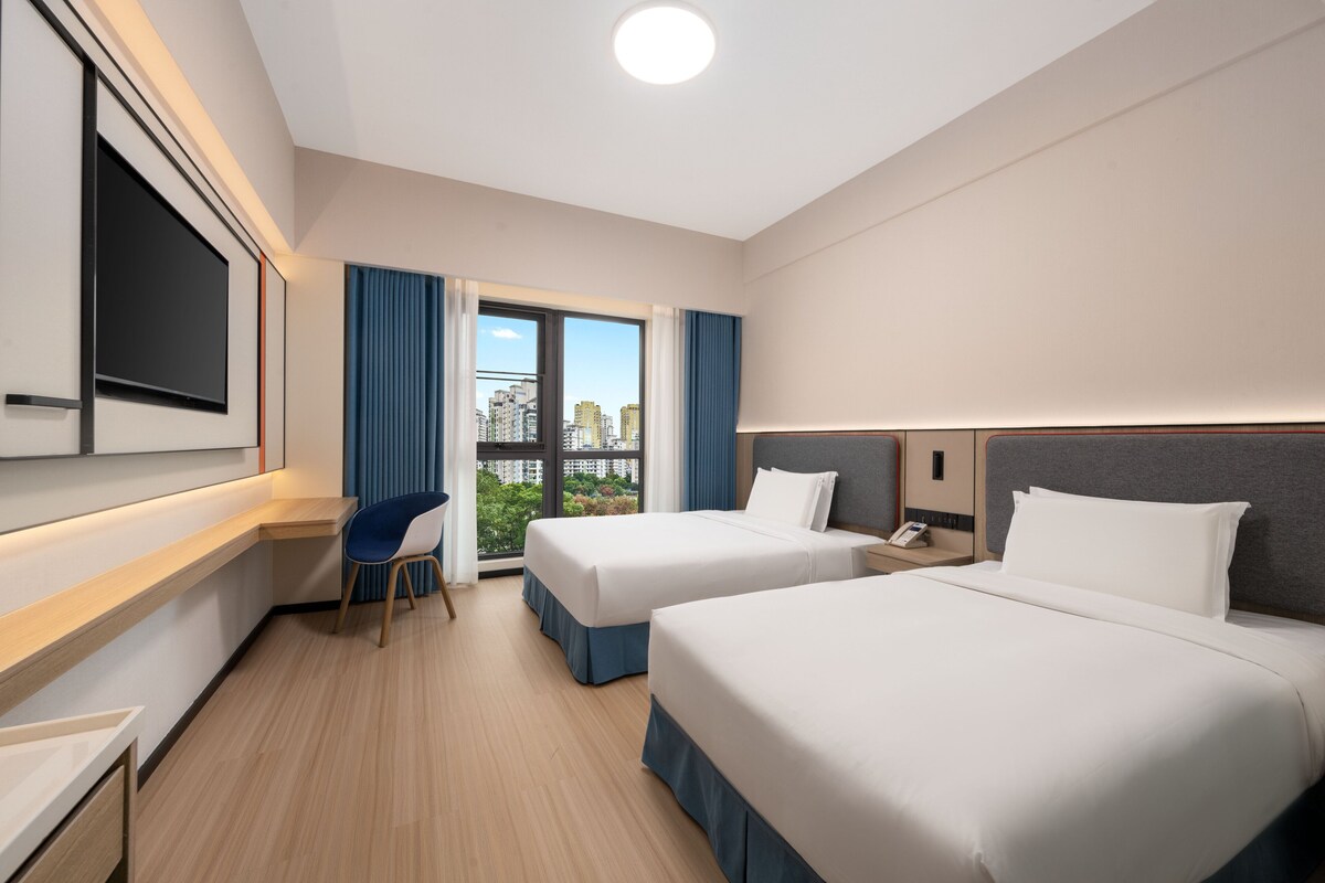 A hotel room with two beds, a desk, a chair, a TV, and a city view.