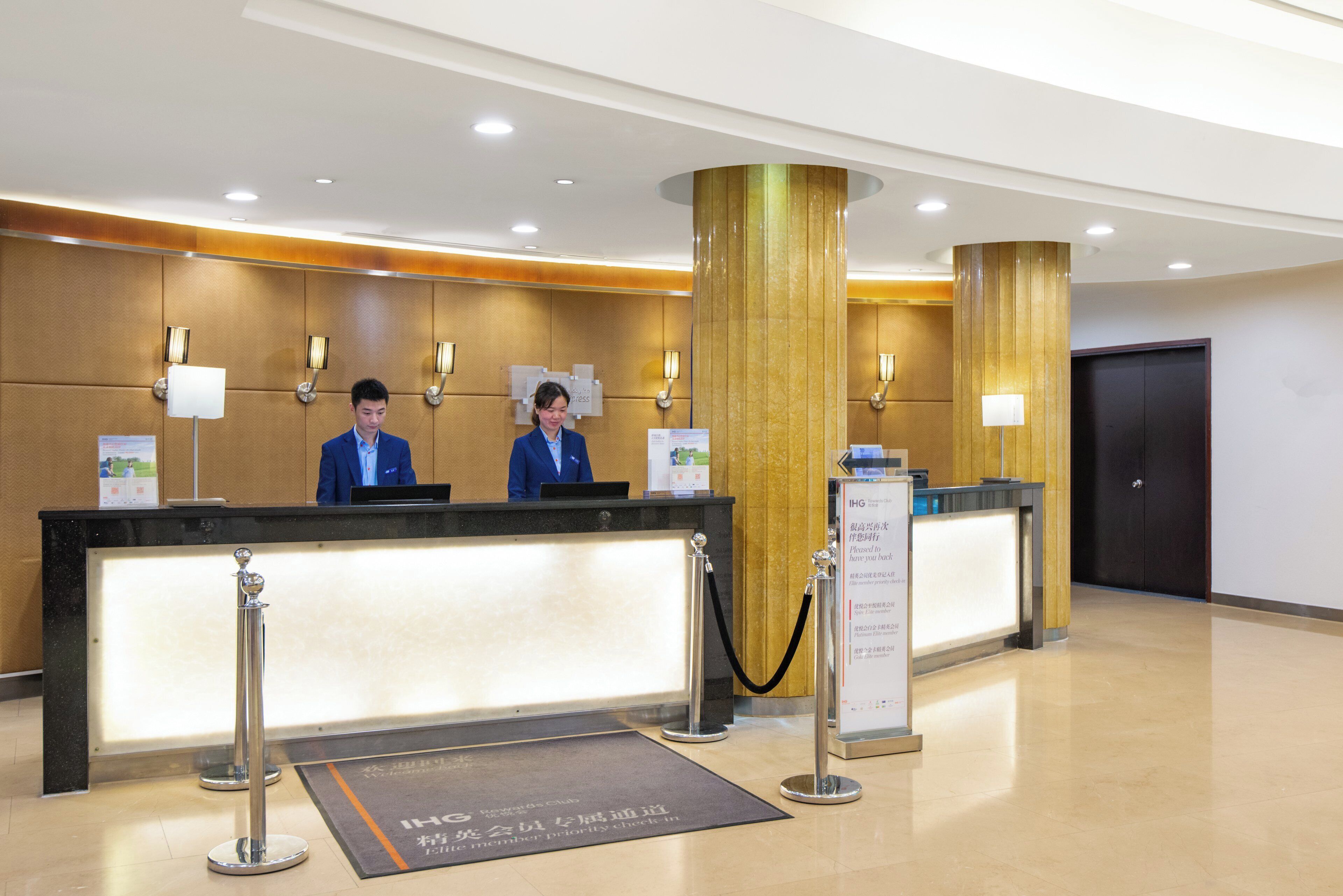 Un mostrador de recepción de hotel con dos empleados, un pilar grande de color dorado y un letrero que indica un área de check-in para miembros.