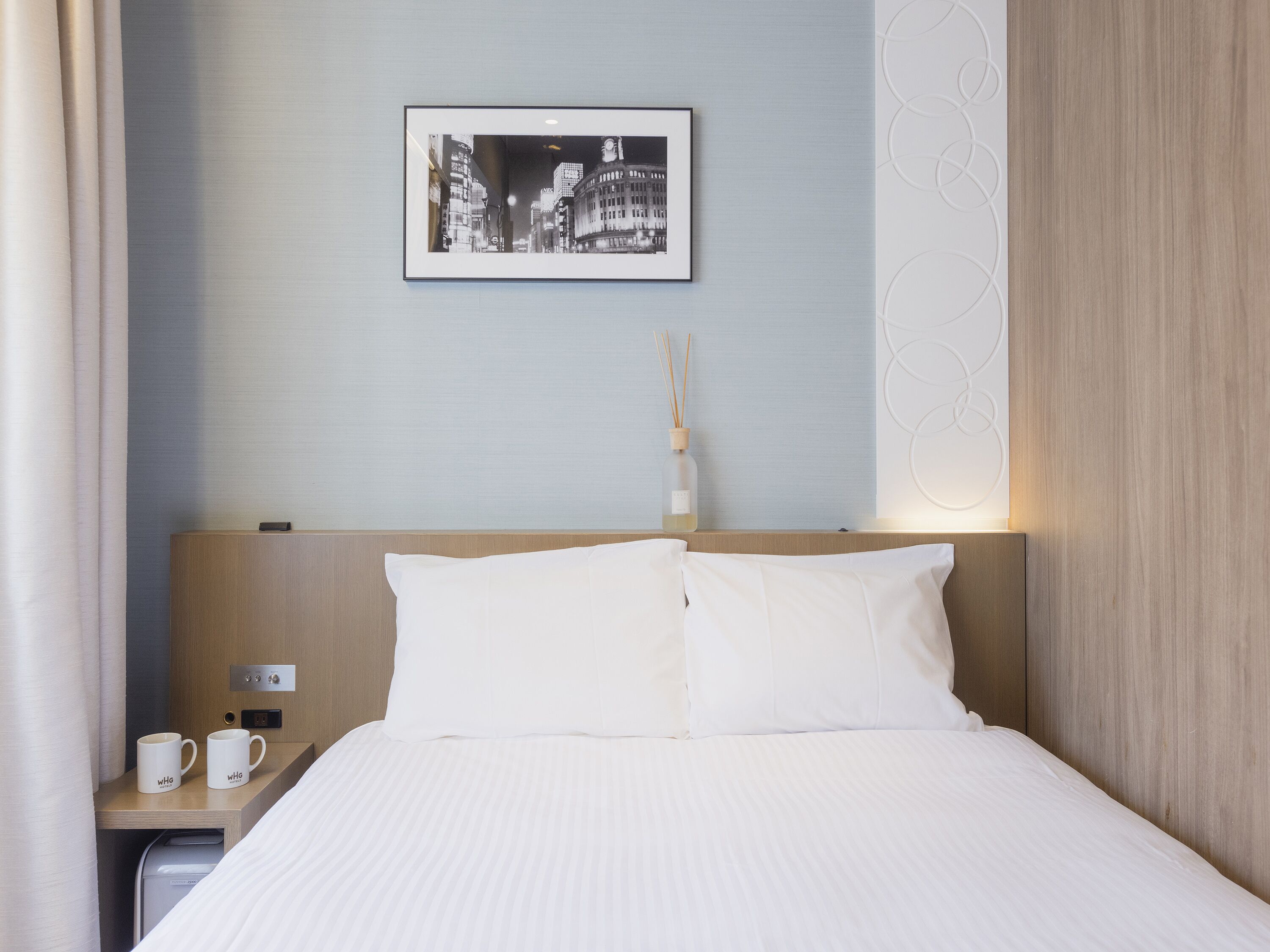 Een netjes opgemaakt bed met witte lakens, een lijst met een stadsgezicht aan de muur, een nachtkastje met twee witte mokken en een flesje reed diffuser.