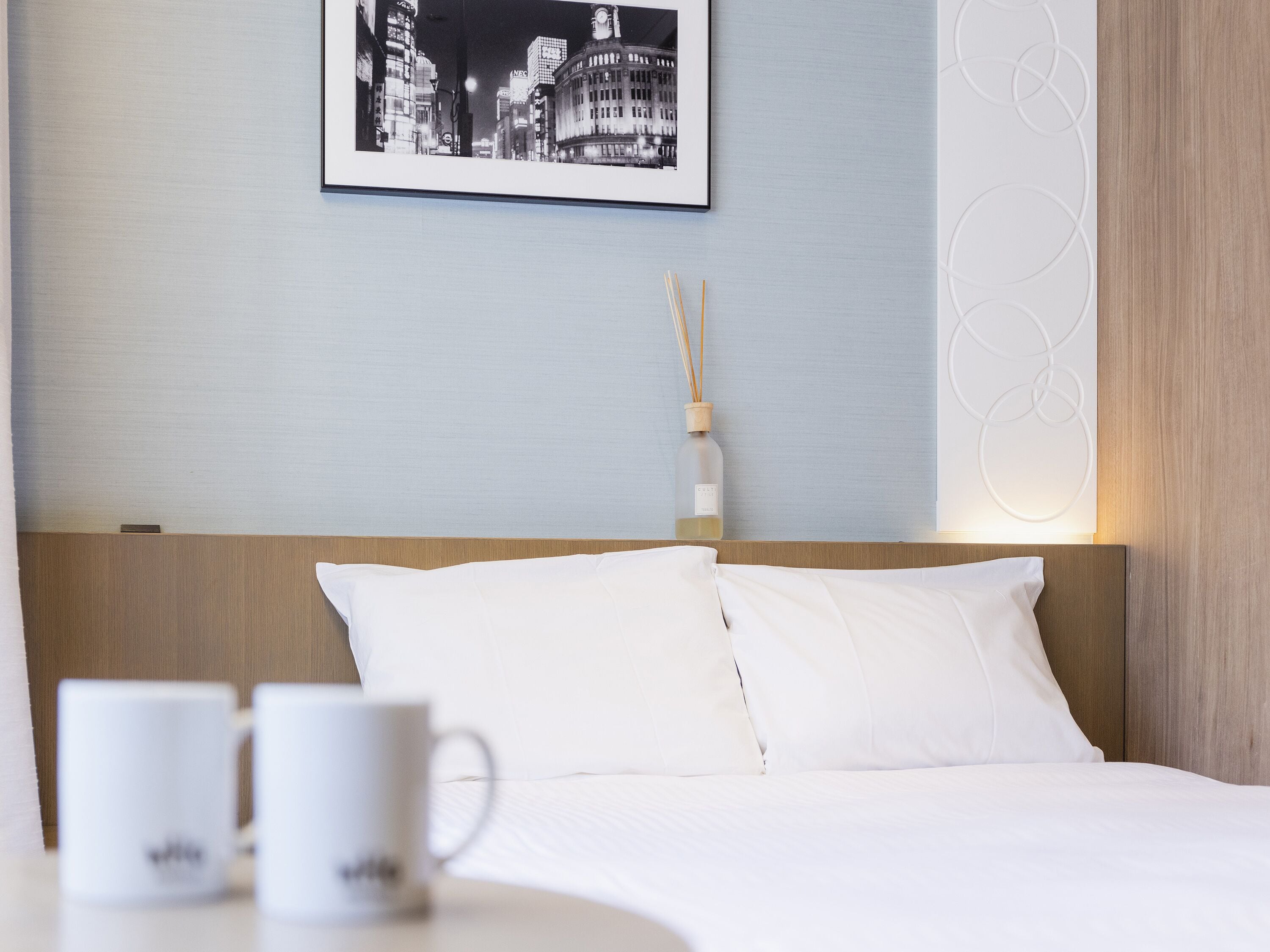 A hotel room with a bed, white pillows, a framed picture of a cityscape, a bottle of reed diffuser, and two white mugs on a table.