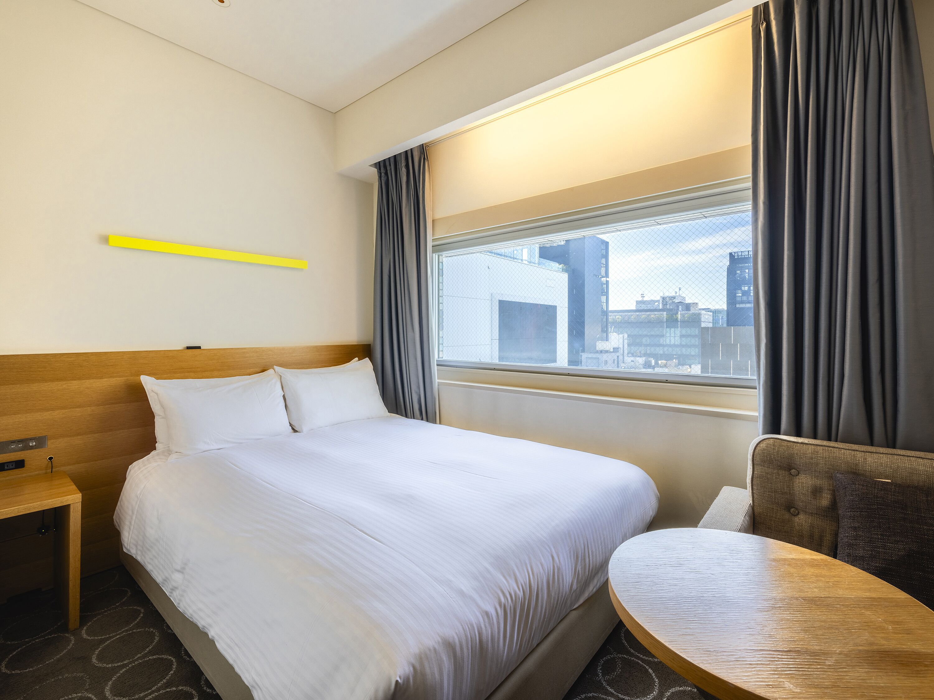 Habitación de hotel con una cama grande, una mesita de noche de madera, una mesa redonda de madera y vistas a la ciudad a través de una ventana con cortinas.