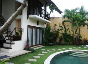 A two-story house with a balcony, a staircase, a swimming pool, and tropical plants.