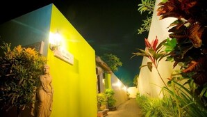 A narrow alleyway with a statue on the left, a brightly lit wall on the right, and potted plants lining the path.