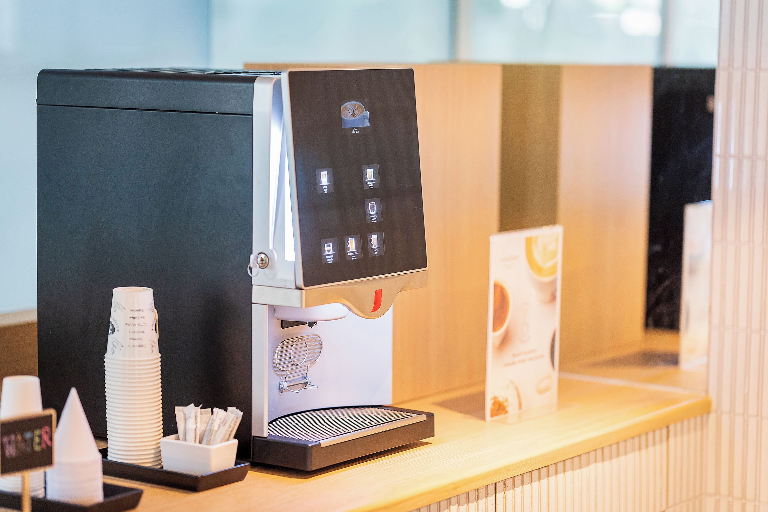 A coffee machine with a digital interface, a stack of disposable cups, and a napkin holder on a counter.