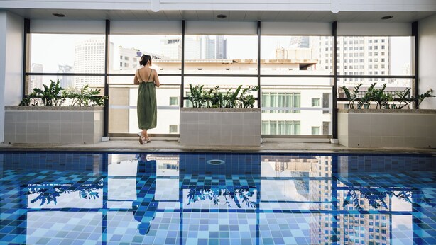 A woman standing by a window overlooking a pool with city buildings in the background. - Citadines Sukhumvit 16 Bangkok (Bangkok)