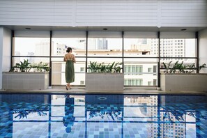 A woman standing by a window overlooking a pool with city buildings in the background. - Citadines Sukhumvit 16 Bangkok (Bangkok)