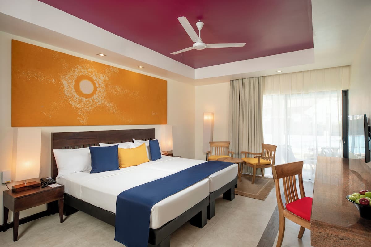 A hotel room with a large bed, a ceiling fan, a dining table with chairs, and a TV.