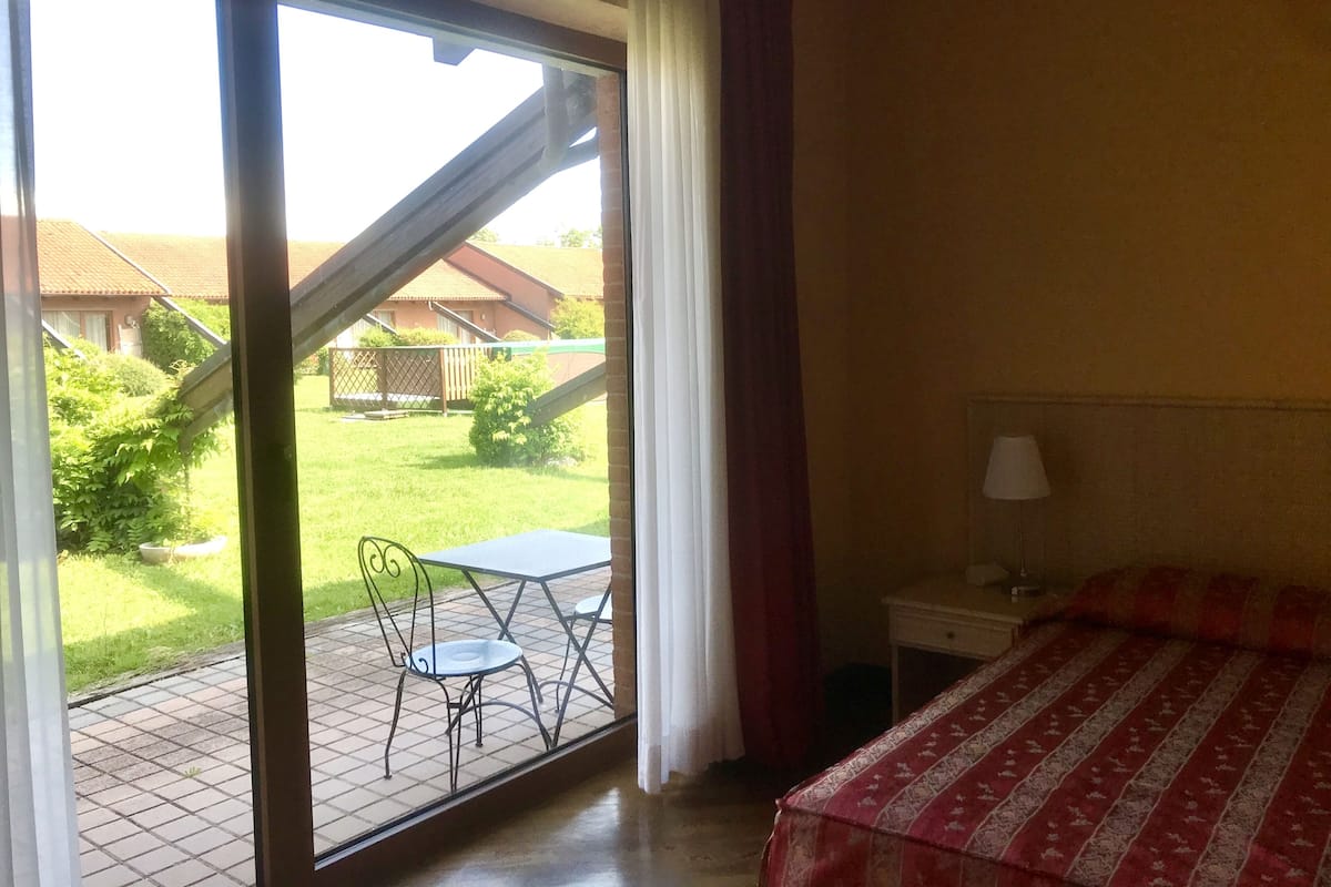 A hotel room with a large sliding glass door leading to a patio with a table and chairs, a bed with a patterned bedspread, and a nightstand with a lamp.