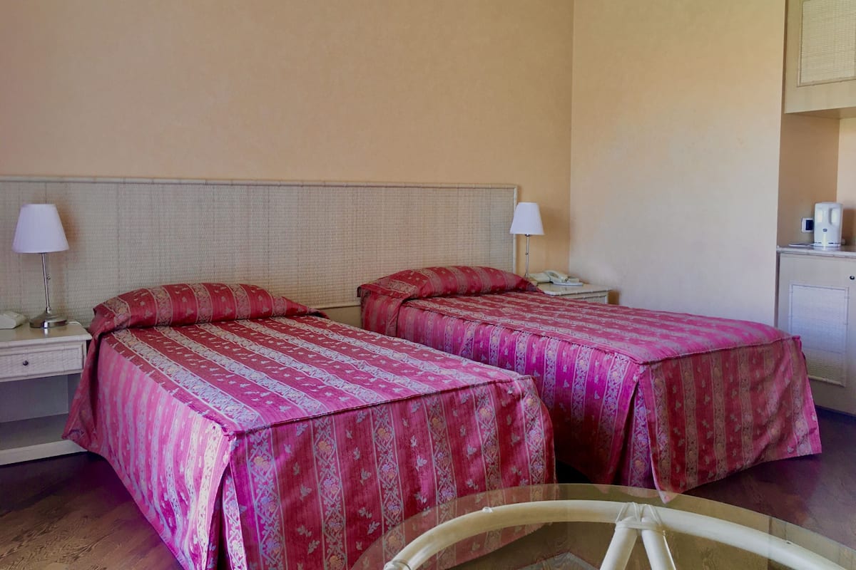 A hotel room with two beds, each with a patterned bedspread, bedside lamps, a nightstand, and a small table with a coffee maker.