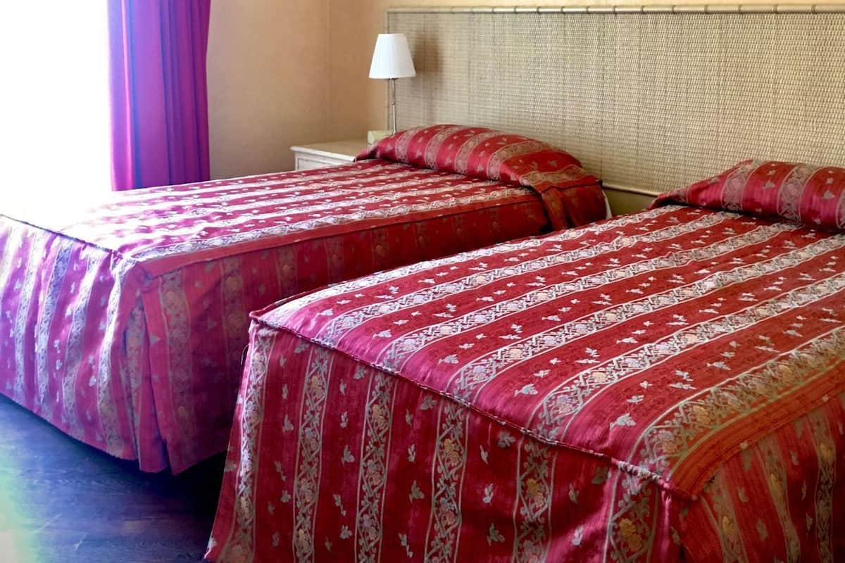 A hotel room with two beds, a nightstand, a lamp, and a window with curtains.