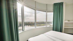 Habitación de hotel con ventana curva, cama y vista a un lago y árboles.