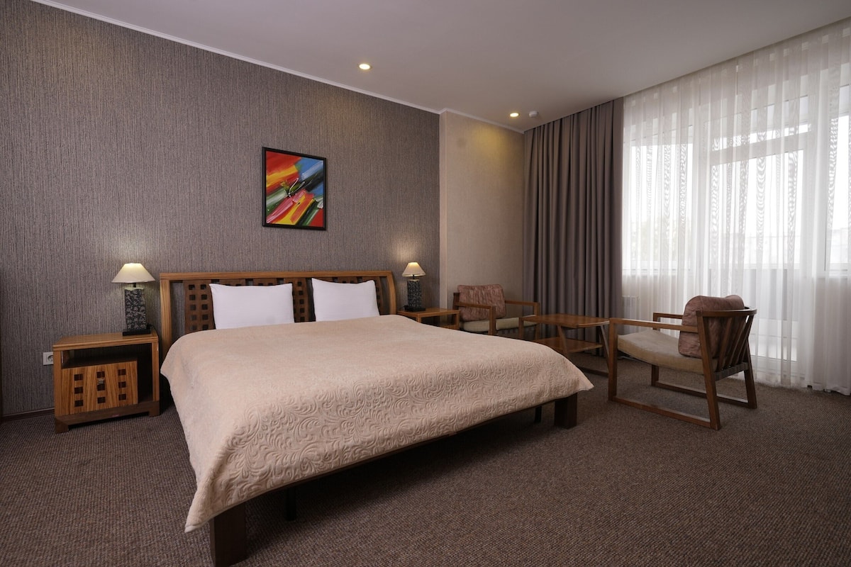 A hotel room with a large bed, two bedside tables, a chair, a desk, and a large window with curtains.