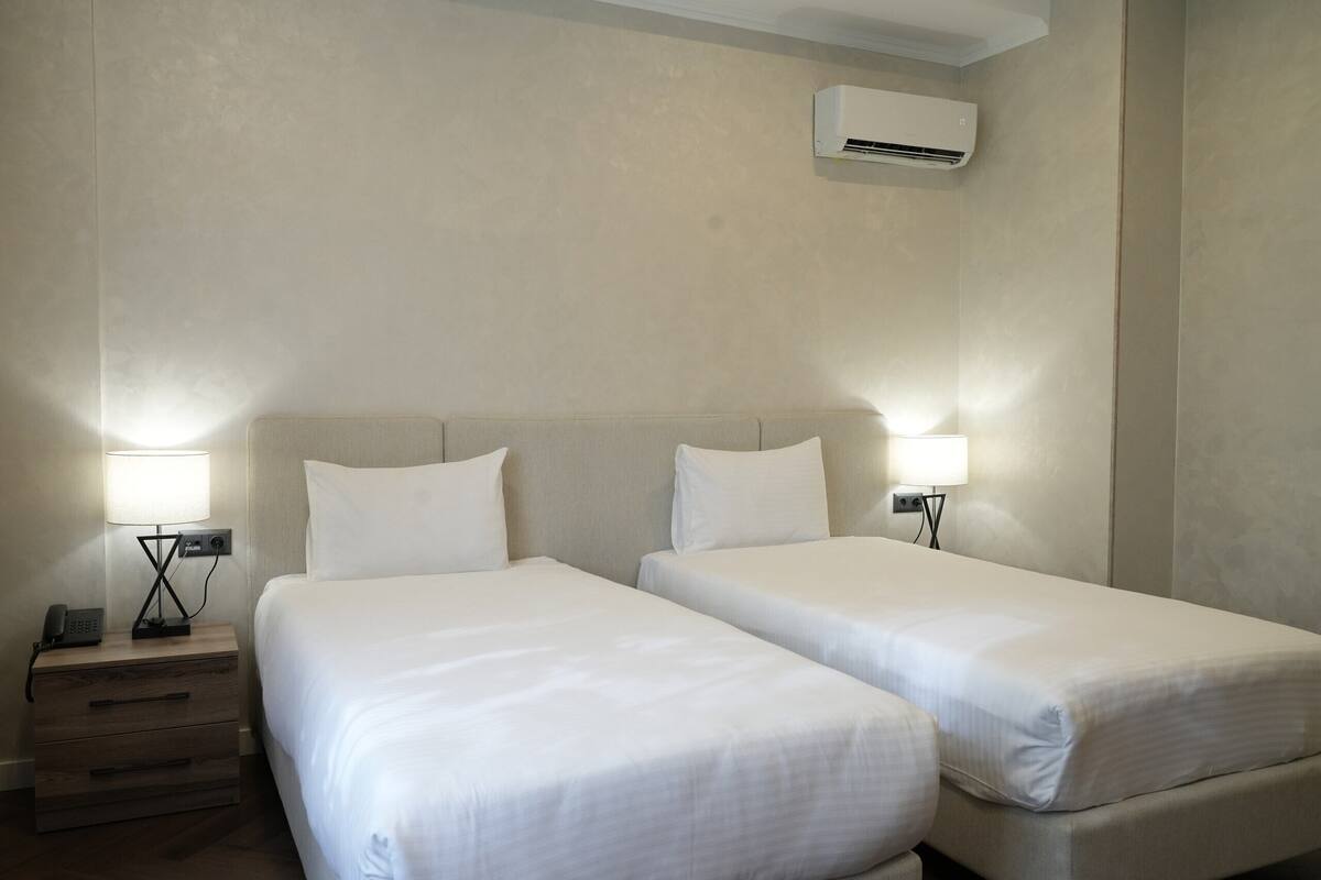 A hotel room with two beds, a bedside table with a lamp, a phone, and an air conditioning unit on the wall.
