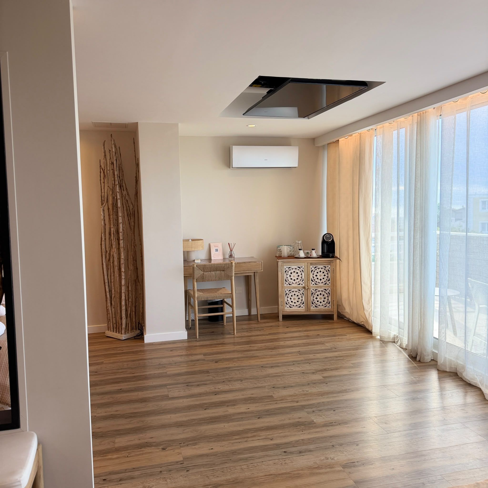 Un'ampia stanza con pavimento in legno, zona pranzo con tavolo e sedie, un armadio con motivi decorativi e un'ampia finestra con tende leggere.