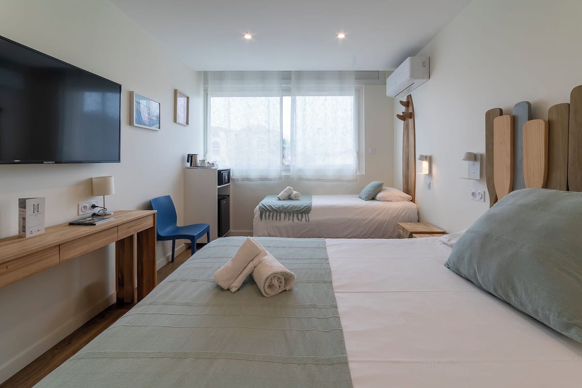 A hotel room with two beds, a desk with a chair, a TV, and a window with sheer curtains.