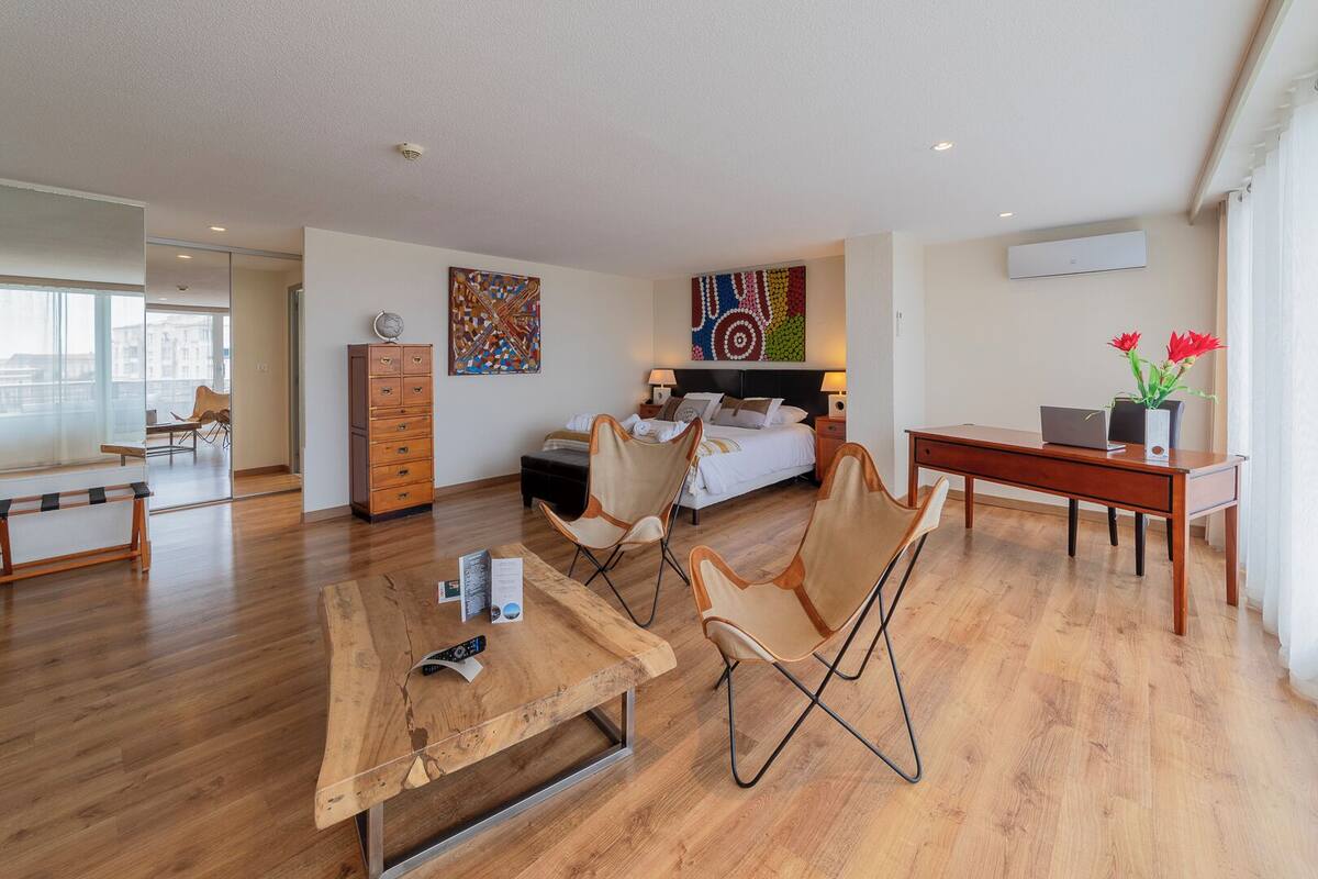 A modern living room with a wooden coffee table, two armchairs, a sofa, and a bed with a colorful abstract painting above it.