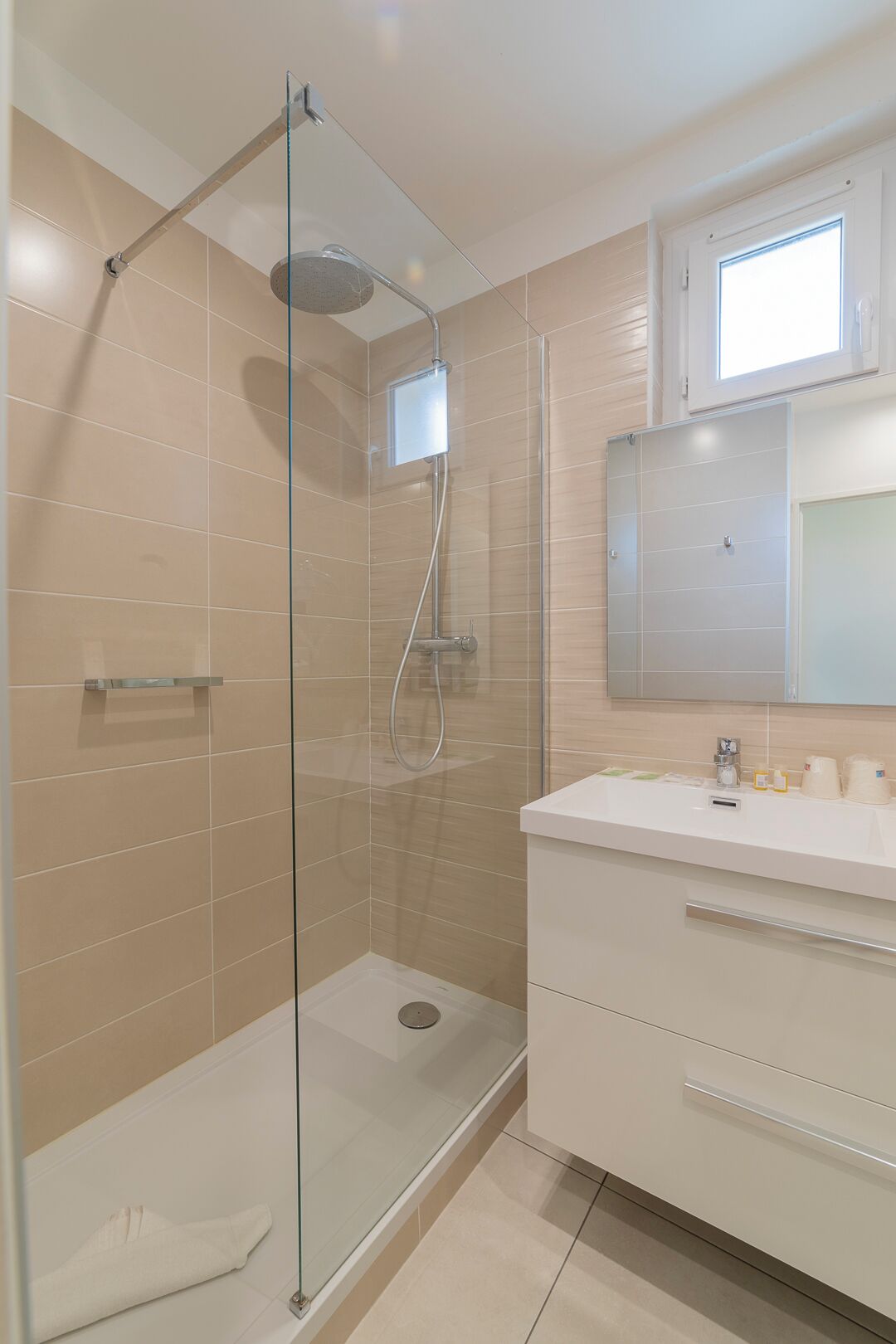 A modern bathroom with a glass-enclosed shower, a white vanity with a mirror, and beige tiled walls.