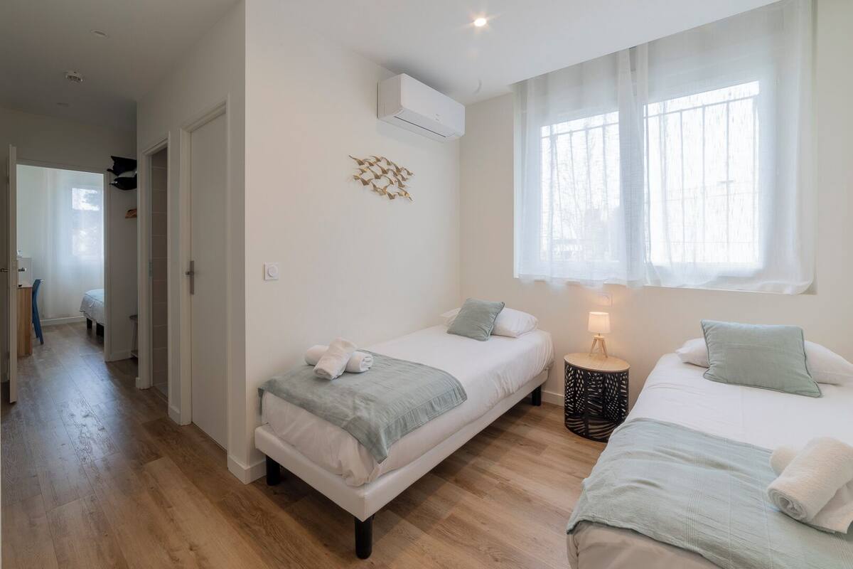 A bedroom with two beds, a bedside table with a lamp, and a window with sheer curtains.