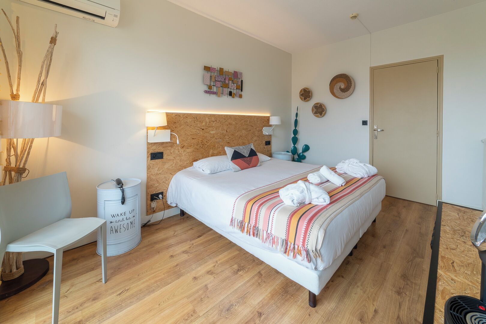 Quarto moderno com cabeceira de madeira, cama com roupa de cama branca e um cobertor colorido, uma cadeira branca, uma mesinha de cabeceira e uma luminária fixada na parede.