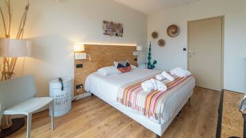 Quarto moderno com cabeceira de madeira, cama com roupa de cama branca e um cobertor colorido, uma cadeira branca, uma mesinha de cabeceira e uma luminária fixada na parede.