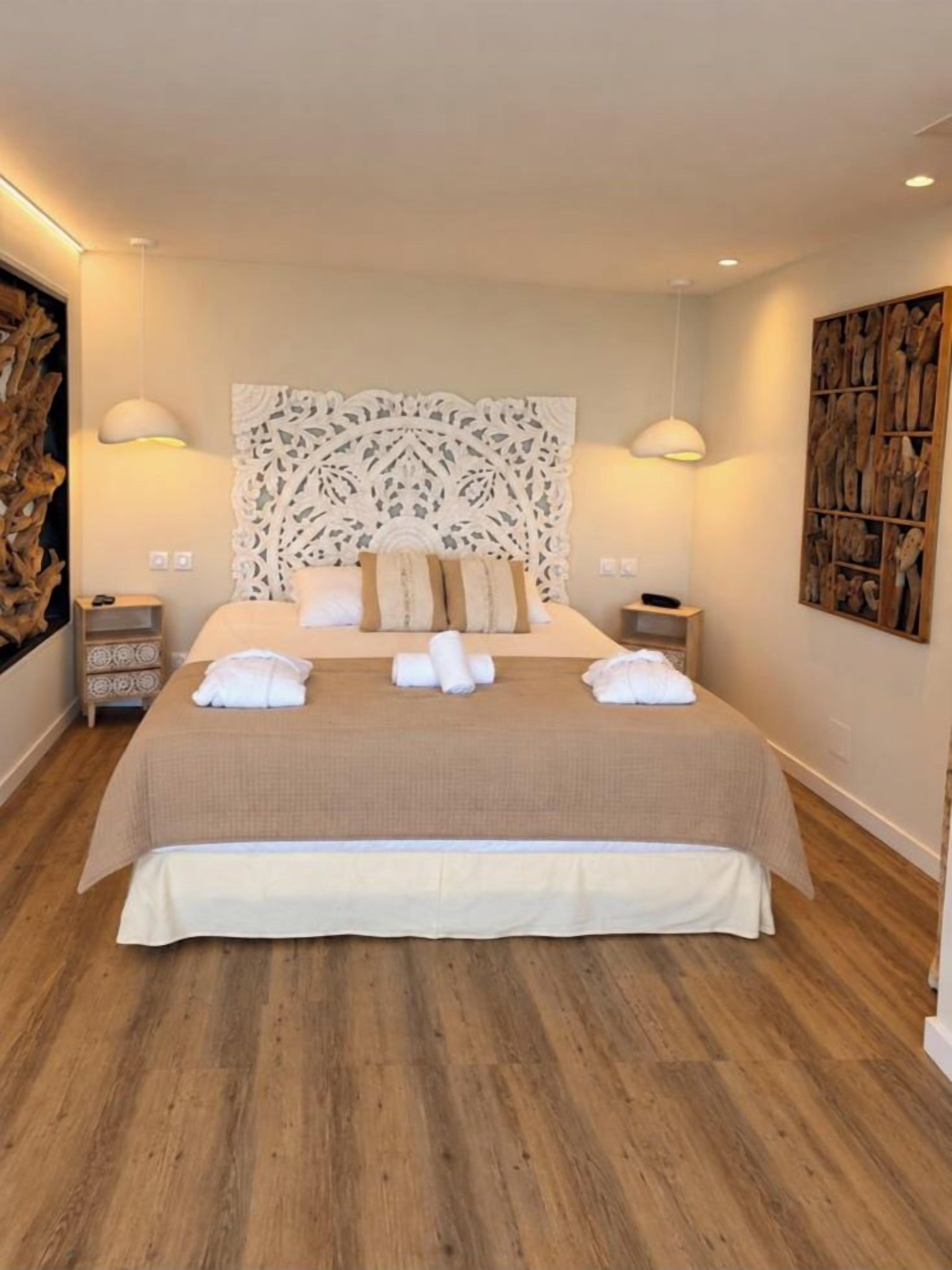 Una cama bien hecha con cubrecama color beige y almohadas blancas, un cabecero decorativo y dos mesitas de noche con lámparas.