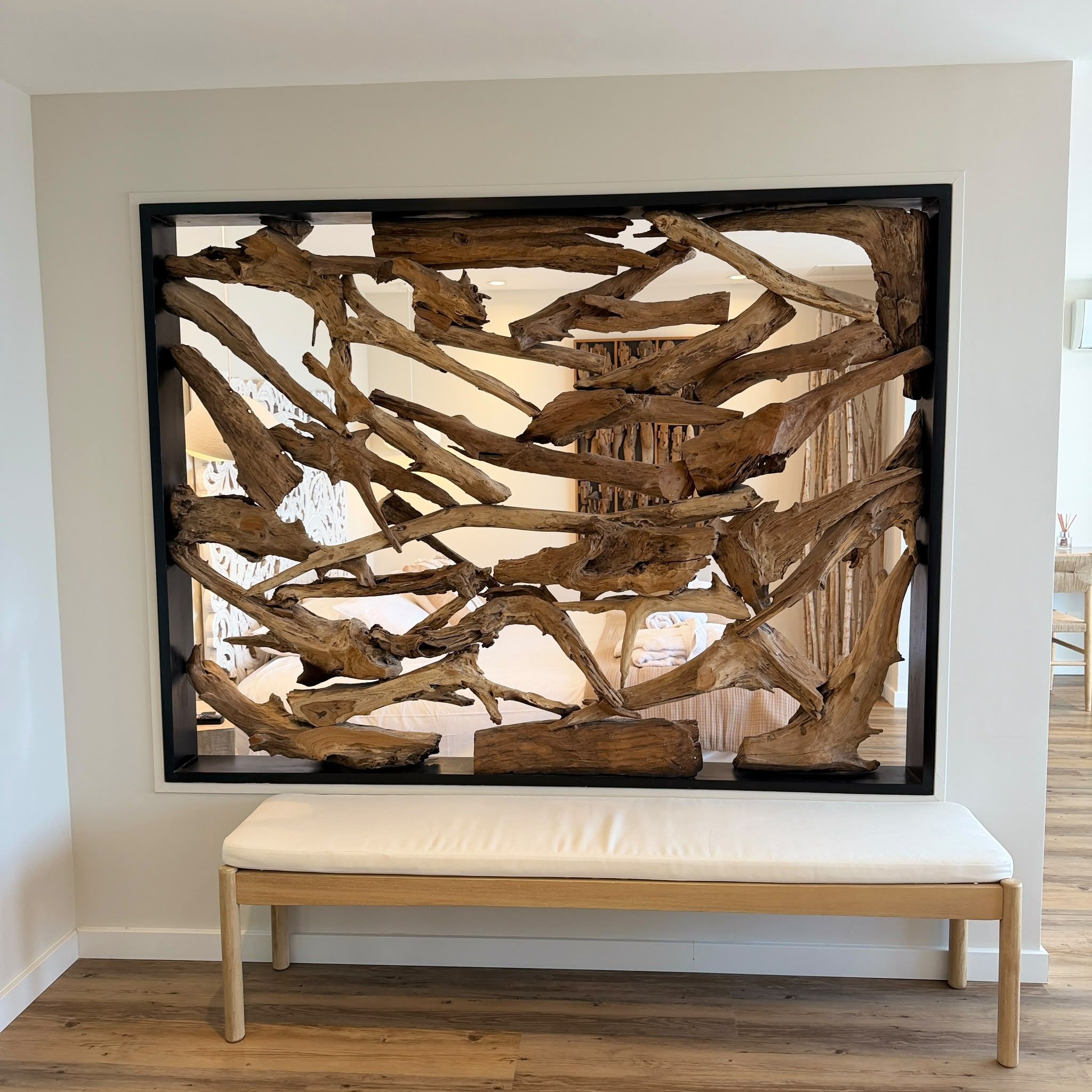 A room with a wooden art installation, a white bench, and a view of a bed with a white headboard.