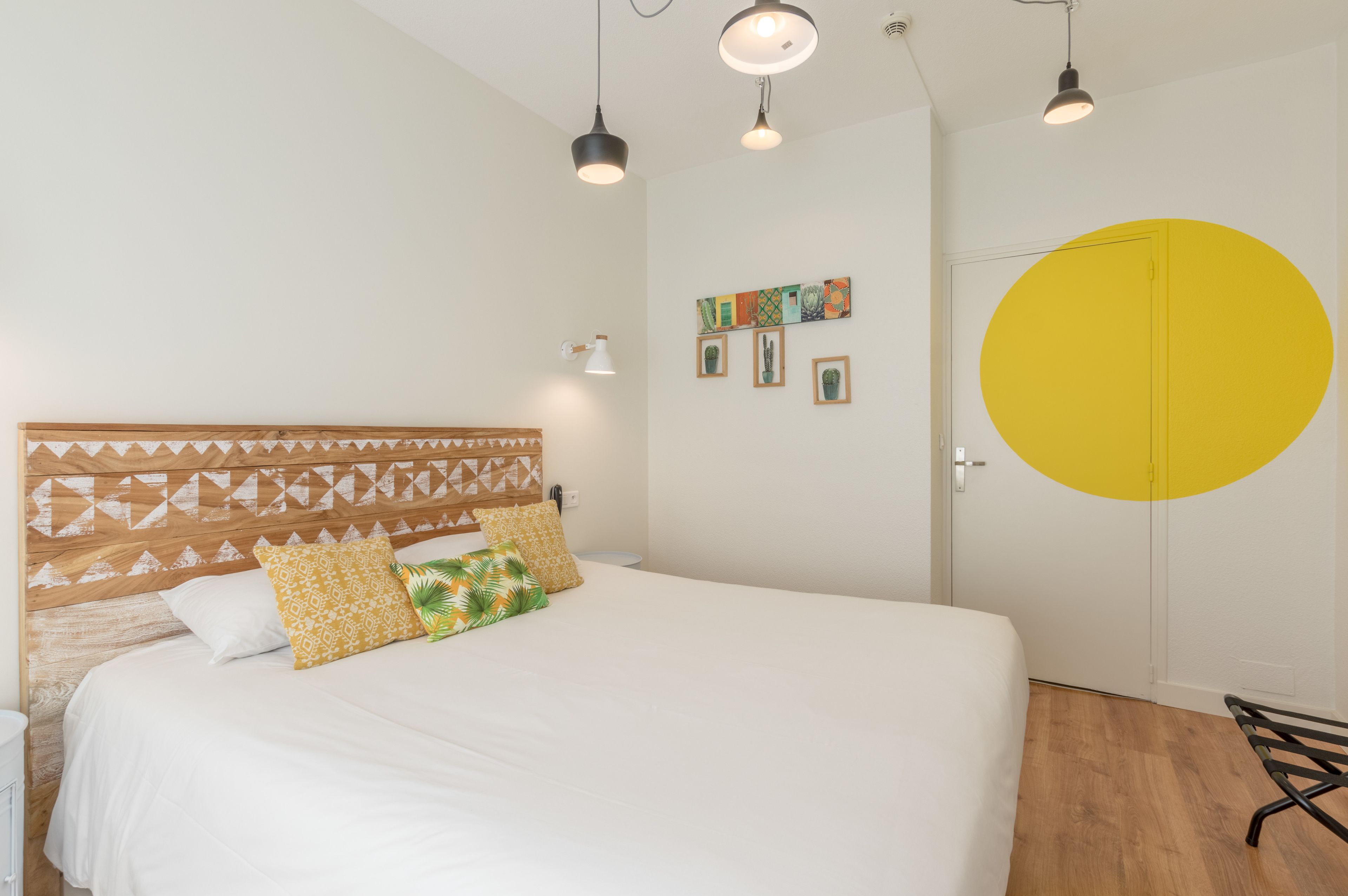 A bedroom with a bed, wooden headboard, multiple pillows, wall-mounted artwork, a door with a large yellow circle, and a bench.