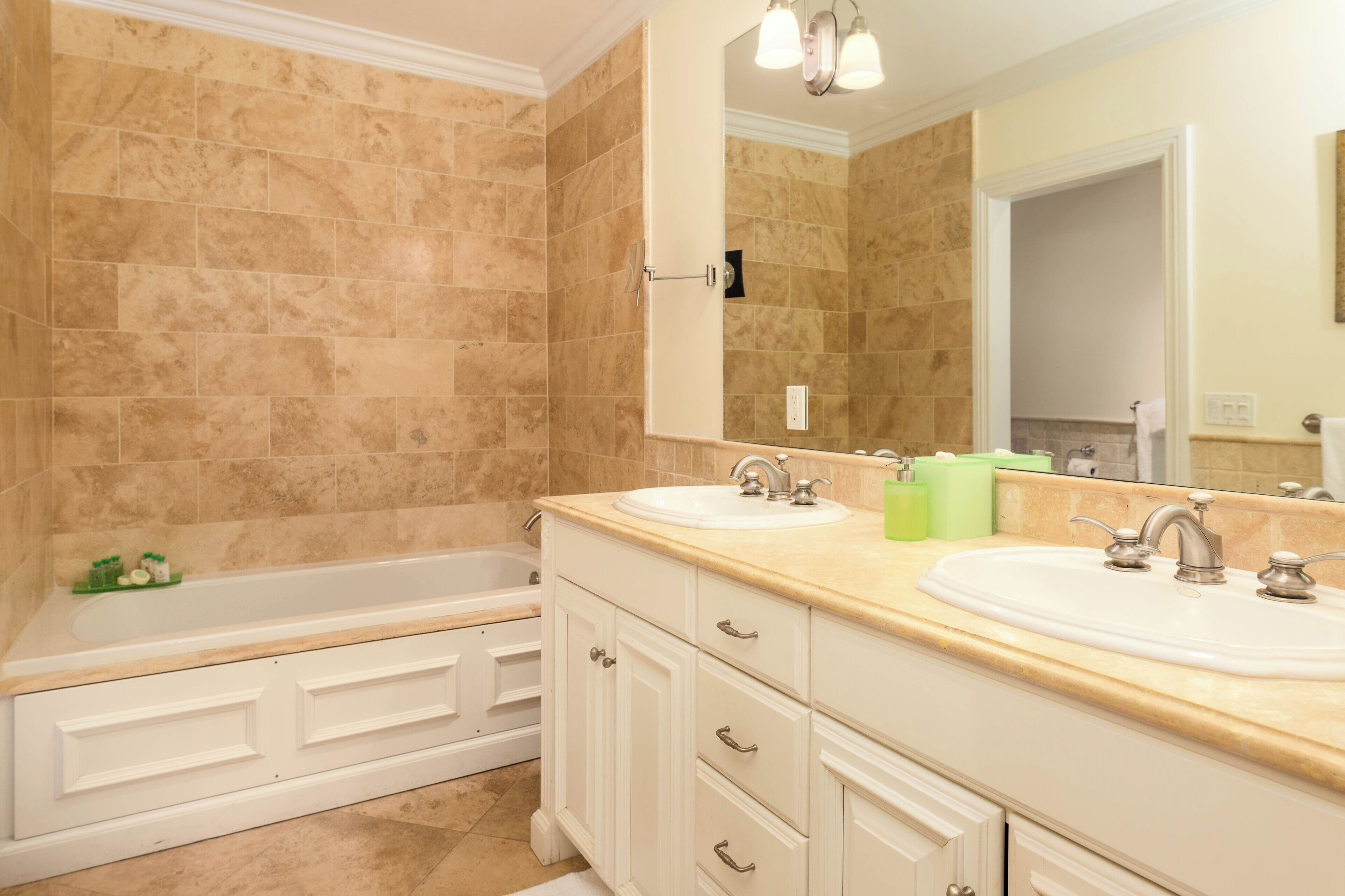 A bathroom with beige tiled walls, a white bathtub, two sinks, and a large mirror above the sink.