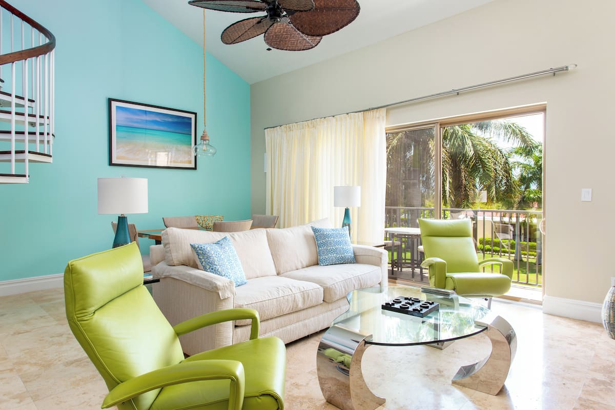 Un salon avec un mur turquoise, un canapé blanc, des fauteuils verts, une table basse en verre et un balcon offrant une vue sur des palmiers.