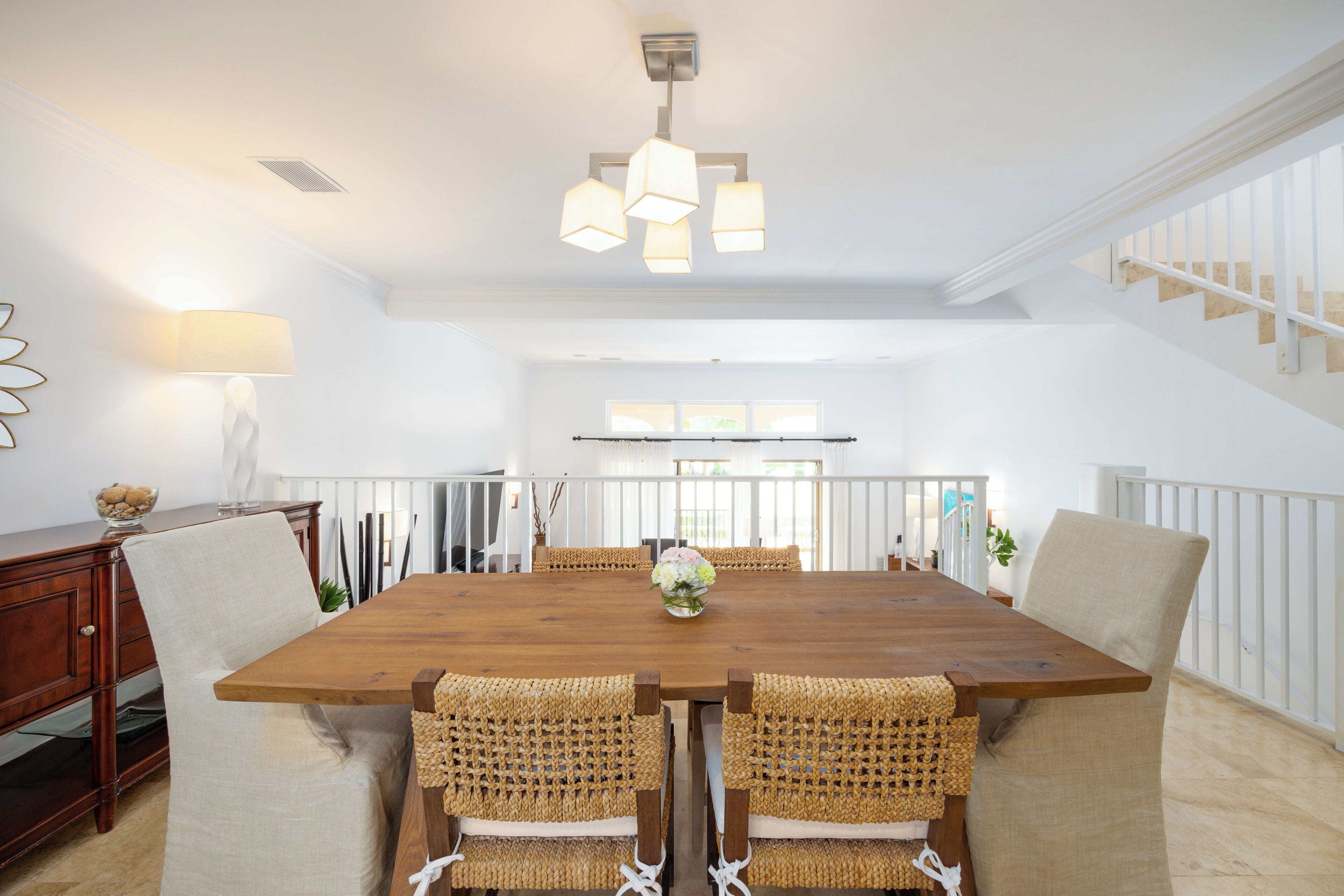 A spacious dining area with a wooden table and woven chairs, a hanging light fixture, and a view of a staircase.