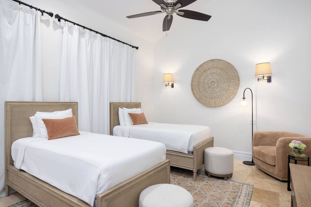 Une chambre d’hôtel avec deux lits simples, un ventilateur de plafond, un élément décoratif rond fixé au mur, deux lampes murales, une lampe sur pied, un pouf rond et un fauteuil marron.