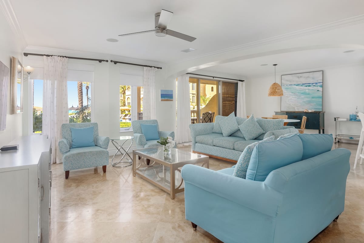 Un salon spacieux avec un grand canapé bleu clair, un fauteuil assorti et une table basse. On y trouve un ventilateur de plafond, une grande fenêtre avec des rideaux et un tableau représentant l’océan accroché au mur.