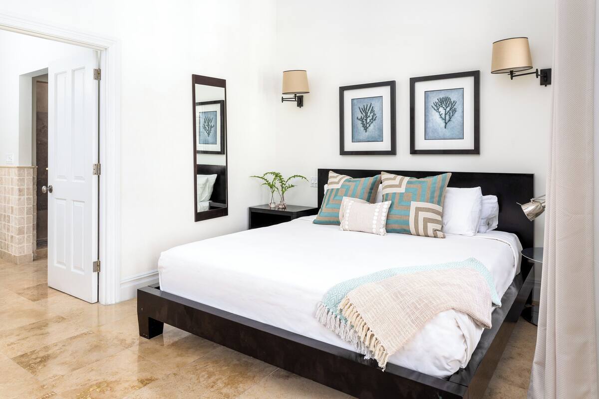 Une chambre à coucher avec un grand lit, deux tables de chevet, une commode, un miroir et des œuvres d’art encadrées accrochées au mur.
