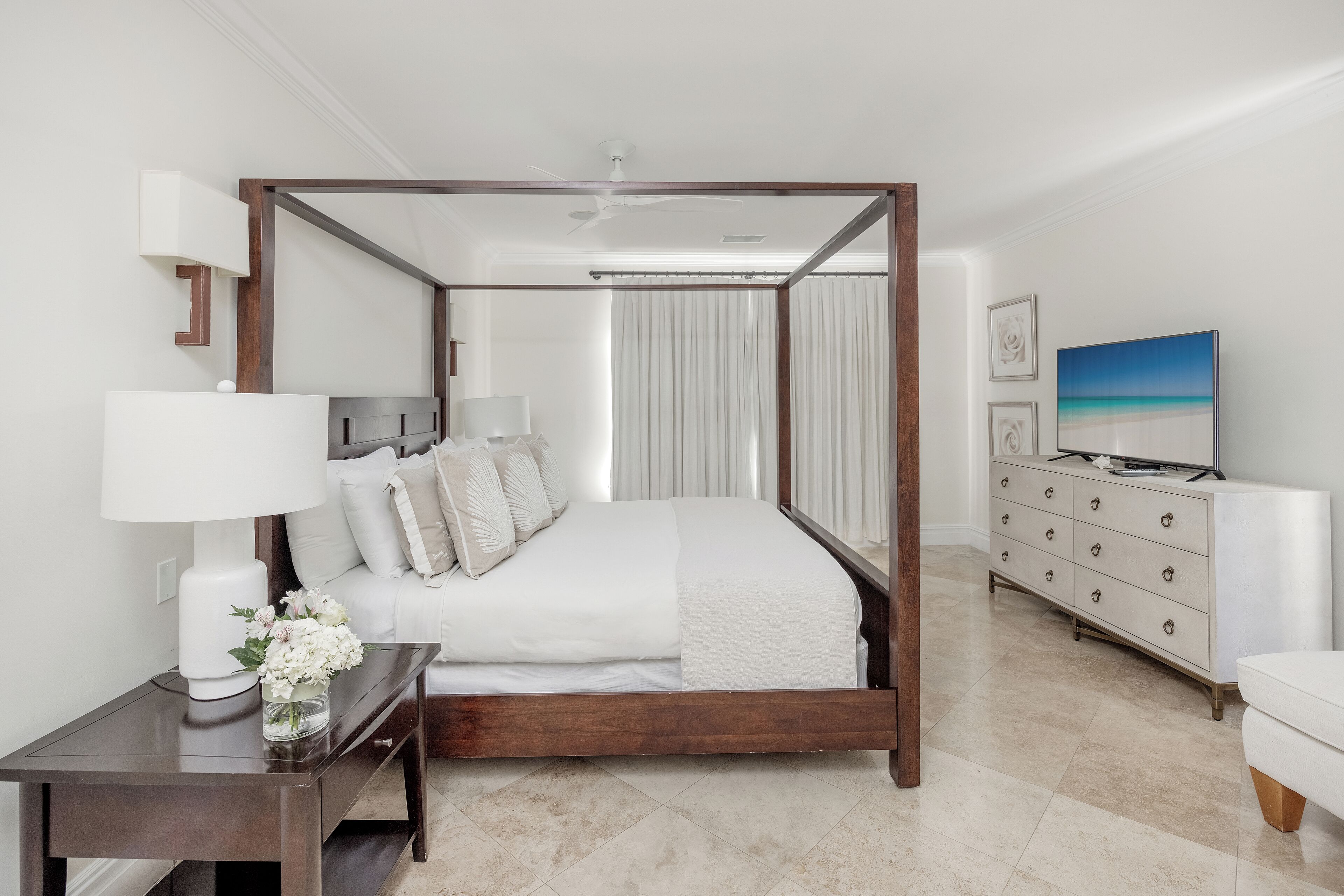 A four-poster bed with a white bedspread, a bedside table with a lamp and flowers, a white dresser with drawers, and a television on the dresser.