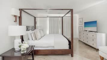 A four-poster bed with a white bedspread, a bedside table with a lamp and flowers, a white dresser with drawers, and a television on the dresser.