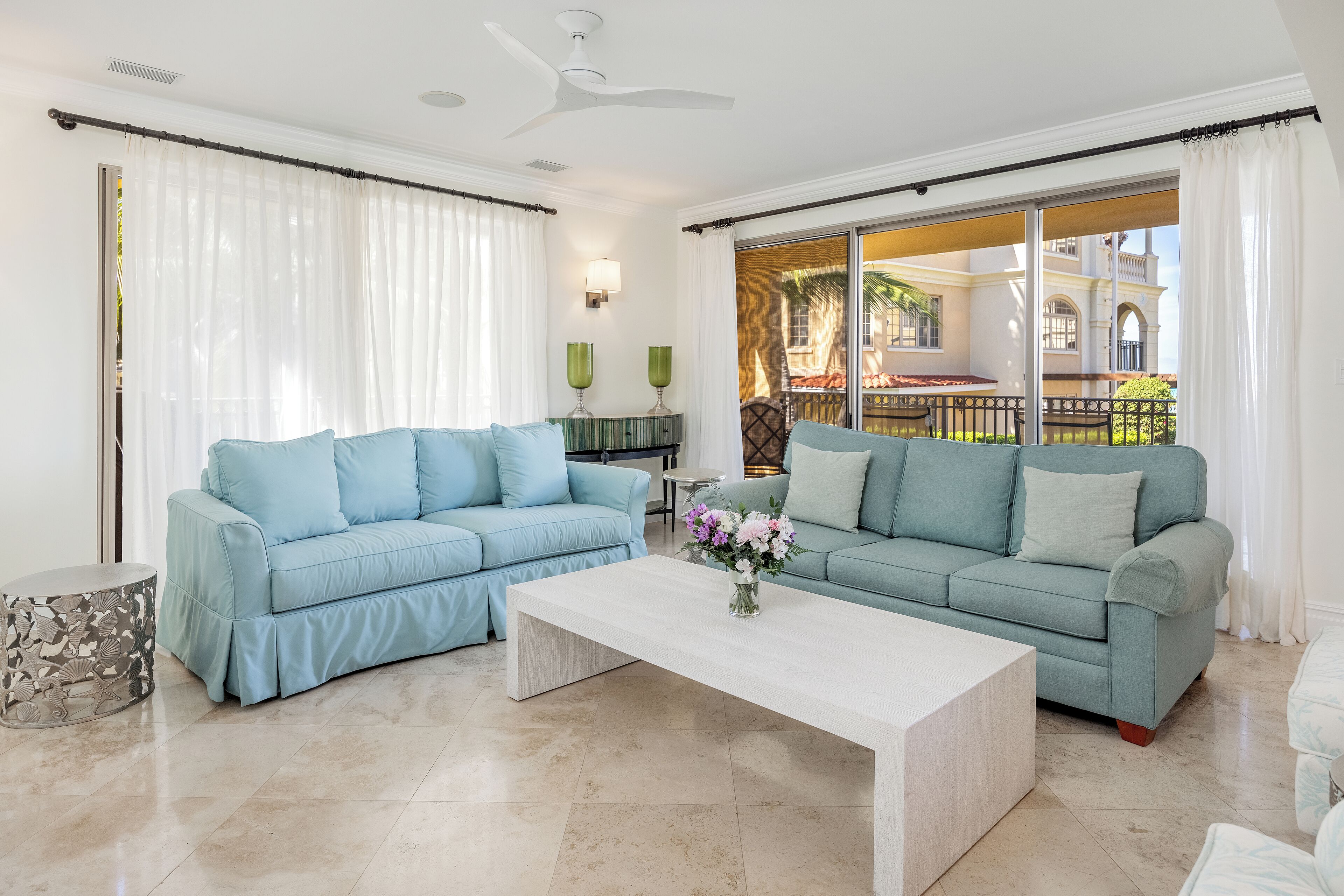 A living room with two light blue sofas, a white coffee table, and a view of a courtyard with a pool and palm trees.