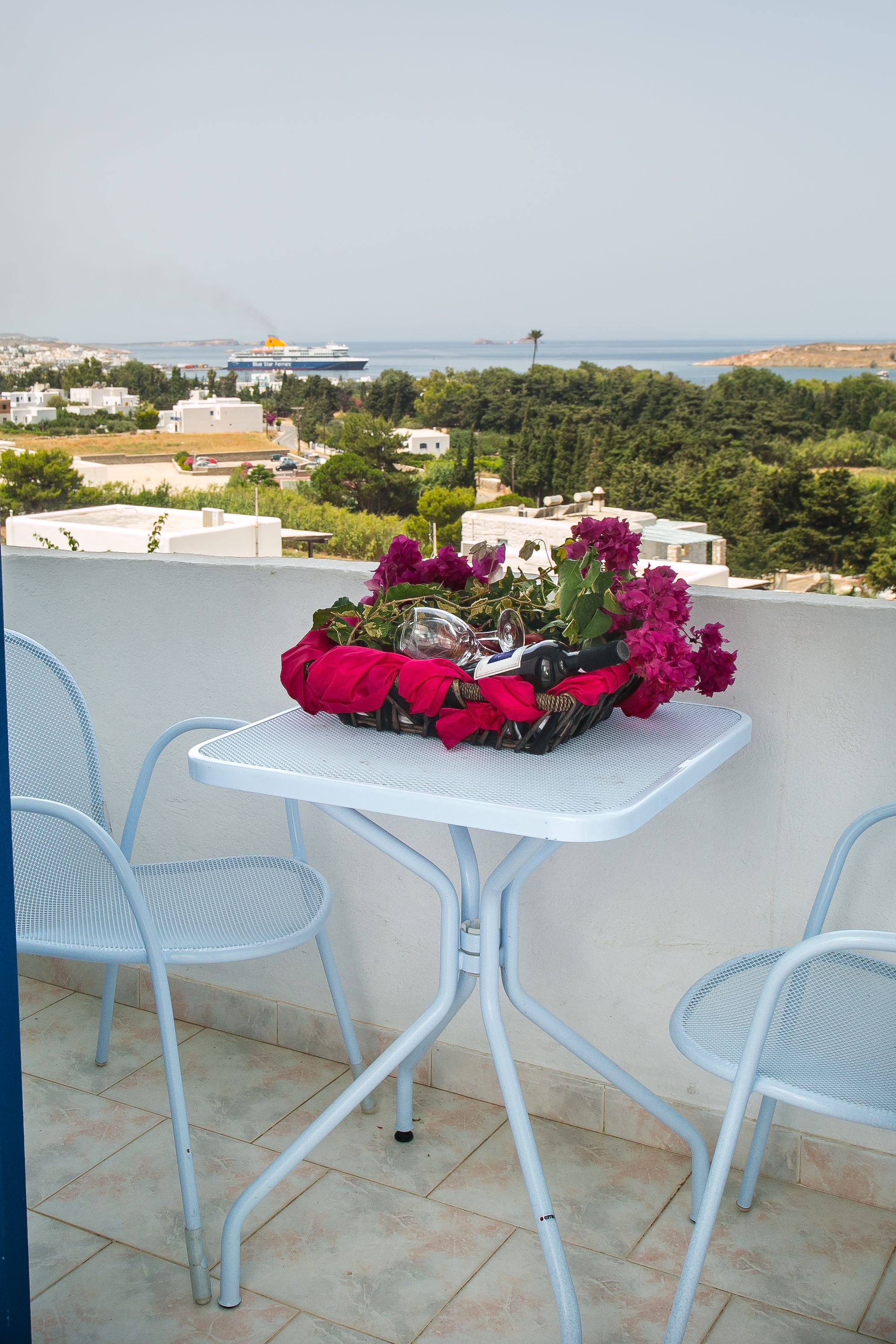 A balcony with a white metal table and chairs, a potted plant with flowers, and a view of the sea and buildings in the background.