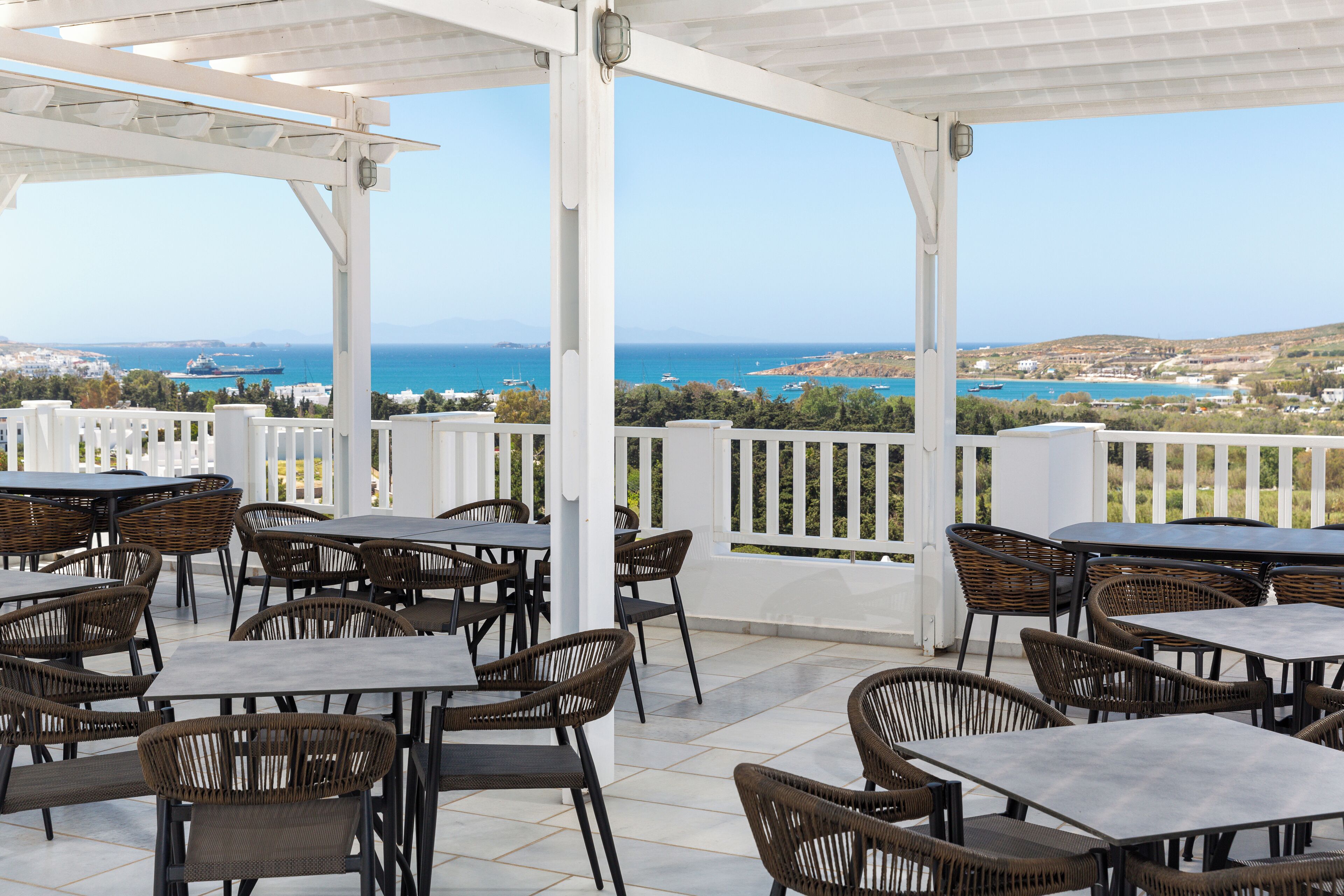 Un patio al aire libre con mesas y sillas de mimbre, con vistas a un paisaje costero con barcos y edificios.