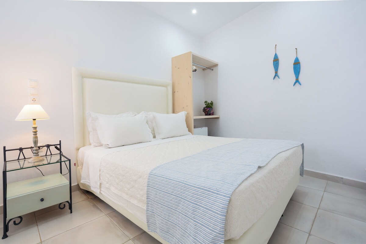 Una cama bien hecha con cabecera blanca, una mesita de noche con lámpara y un estante con una planta.