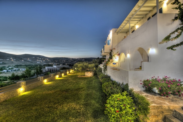 A well-lit outdoor area with a stone pathway, a grassy lawn, and a white building with arches and a balcony.