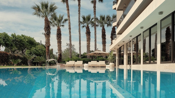 A swimming pool with lounge chairs and palm trees, surrounded by a modern building with large windows. - Golden Sun (Glyfada)