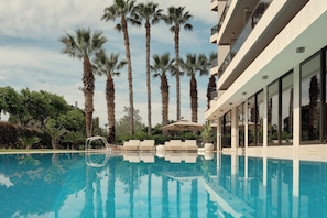 Piscina con sillones de descanso y palmeras, rodeada por un edificio moderno con grandes ventanales.