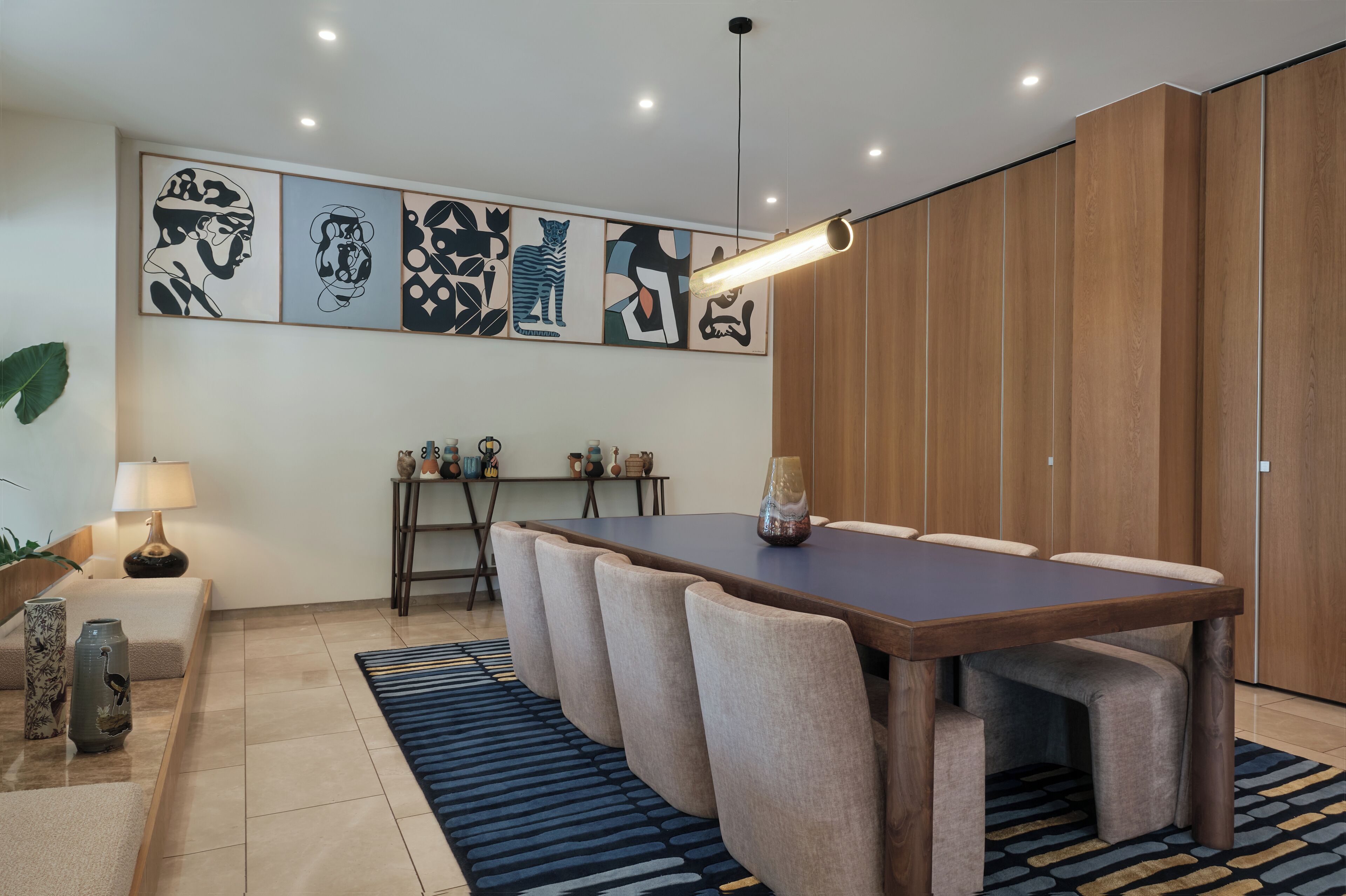 A modern dining area with a long table, cushioned chairs, and a blue patterned rug.