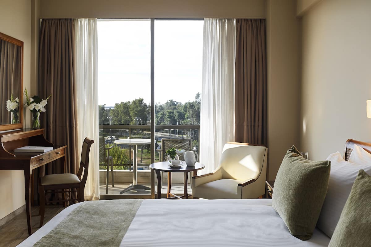A hotel room with a bed, a desk with a chair, a small table with a cup and flowers, and a view of a bridge and greenery through large windows.