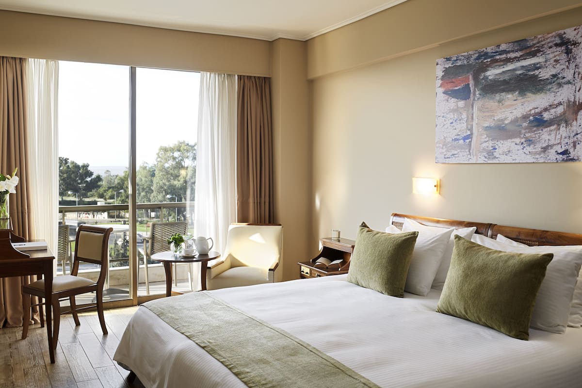 A hotel room with a large bed, a desk, a chair, a small table, and a balcony with a view.